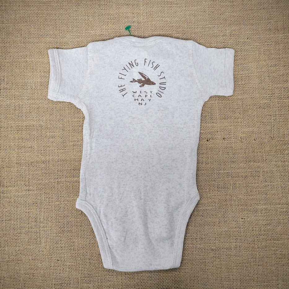 An oatmeal baby onesie featuring a cartoon flying fish. The text "THE FLYING FISH STUDIO, WEST CAPE MAY NJ" is printed around & below the design.
