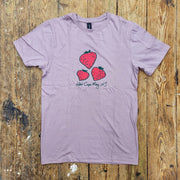 A light purple t-shirt lies flat on a wooden surface, featuring a graphic of three red strawberries of varying sizes. Below the strawberries, "West Cape May, NJ" is handwritten in a casual script.