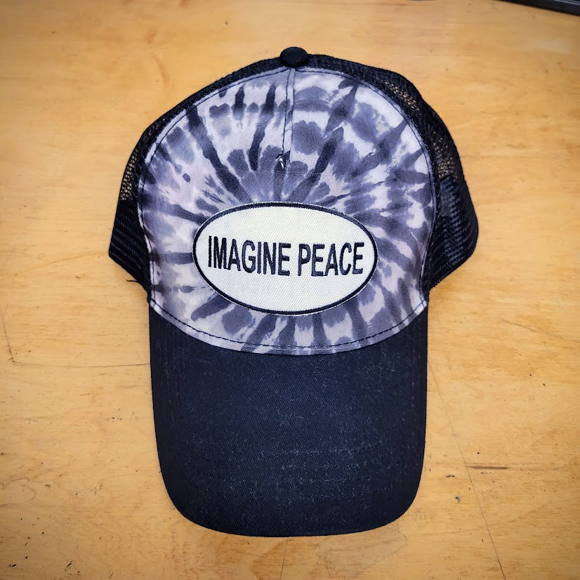 A black tie-dye trucker hat with an 'Imagine Peace' patch on it.