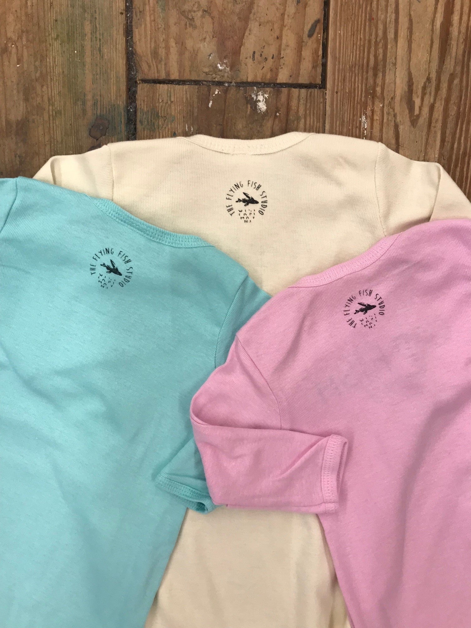 Three sets of layettes in blue, cream and pink featuring the 'Flying Fish' logo on the back neck in black ink.