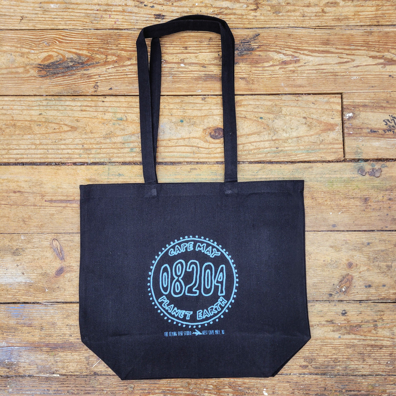 A black tote bag lies flat on a light brown wooden surface. The bag has two handles extending upwards. In the center of the bag, a light blue circular design features the text "CAPE MAY" at the top, "PLANET EARTH" at the bottom, and the number "08204" prominently in the center. Below the circle, smaller text reads "THE FLYING PIG STUDIO WEST CAPE MAY, NJ."