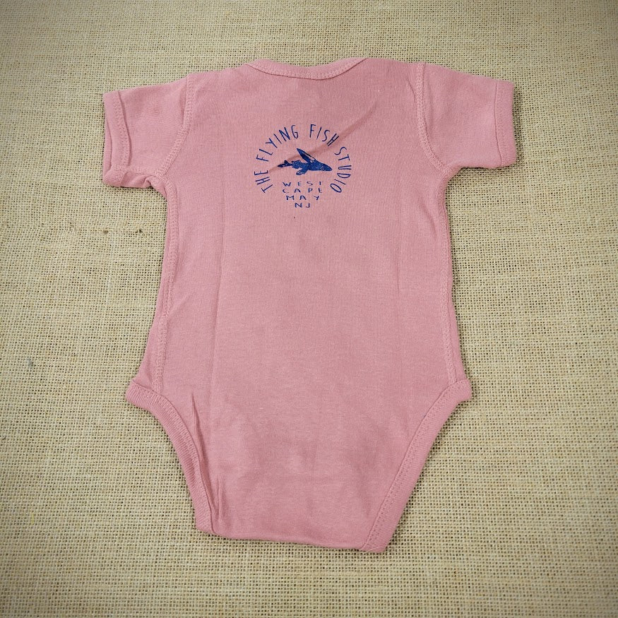 A mauve baby onesie featuring a cartoon flying fish. The text "THE FLYING FISH STUDIO, WEST CAPE MAY NJ" is printed around & below the design.