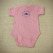 A mauve baby onesie featuring a cartoon flying fish. The text "THE FLYING FISH STUDIO, WEST CAPE MAY NJ" is printed around & below the design.