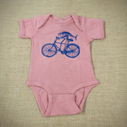 A mauve baby onesie with a graphic of a fish riding a bike.