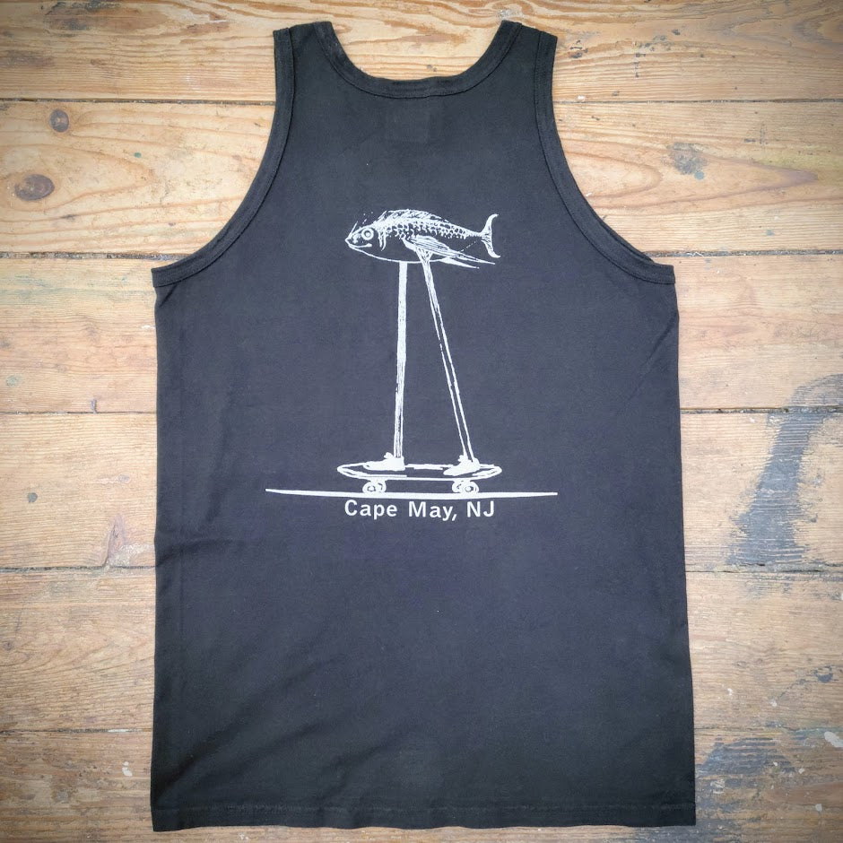Black tank top with a design of a fish riding on a skateboard on the back in silver ink, laid flat on a wooden surface.