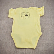 A butter yellow baby onesie featuring a cartoon flying fish. The text "THE FLYING FISH STUDIO, WEST CAPE MAY NJ" is printed around & below the design.