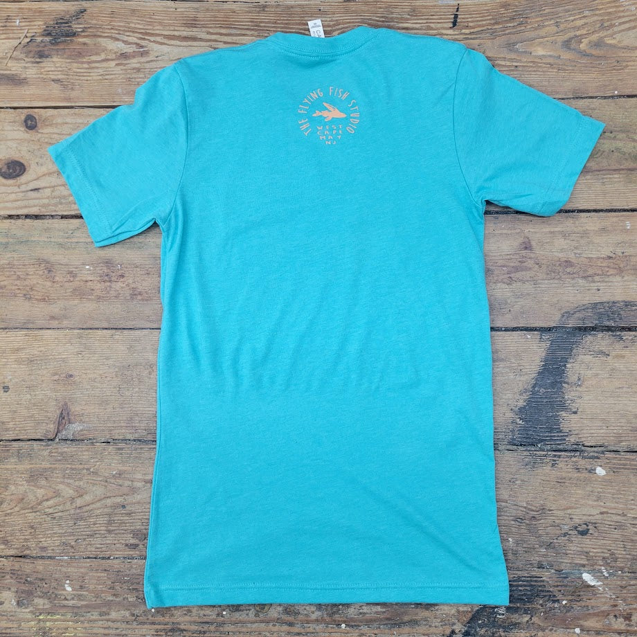 A turquoise t-shirt lies flat on a weathered wooden surface. On the upper back of the shirt, there's a light orange graphic. The graphic is circular and contains the text "THE FLYING FISH STUDIO" in a circular arrangement, with the words "WEST CAPE MAY NJ" underneath. The wooden planks beneath the shirt are light brown with visible grain and some darker markings. A white tag is visible at the neckline of the t-shirt.

