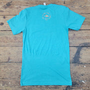 A turquoise t-shirt lies flat on a weathered wooden surface. On the upper back of the shirt, there's a light orange graphic. The graphic is circular and contains the text "THE FLYING FISH STUDIO" in a circular arrangement, with the words "WEST CAPE MAY NJ" underneath. The wooden planks beneath the shirt are light brown with visible grain and some darker markings. A white tag is visible at the neckline of the t-shirt.
