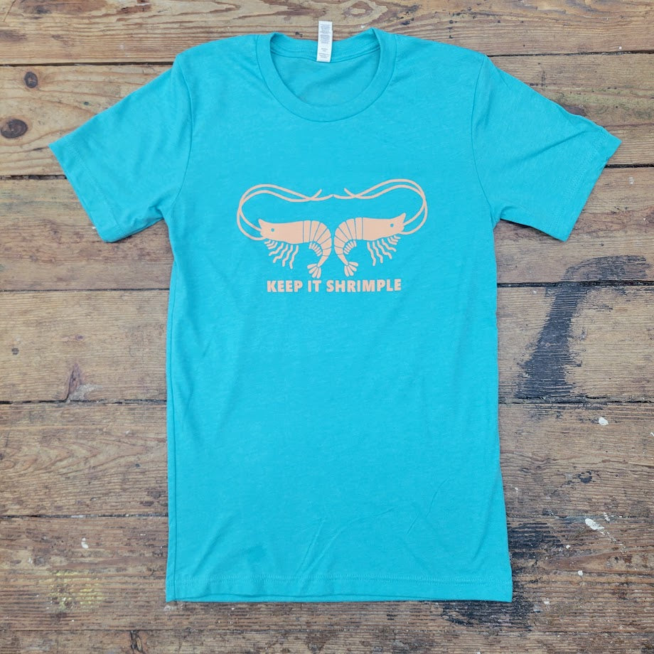 A turquoise t-shirt lies flat on a weathered wooden surface. The shirt features a graphic in a light orange color depicting two stylized shrimp facing away from each other, with their antennae curving upwards and outwards to almost touch. Below the shrimp, in a simple sans-serif font, are the words "KEEP IT SHRIMPLE". The wooden planks beneath the shirt are light brown with visible grain and some darker markings. A white tag is visible at the neckline of the t-shirt.