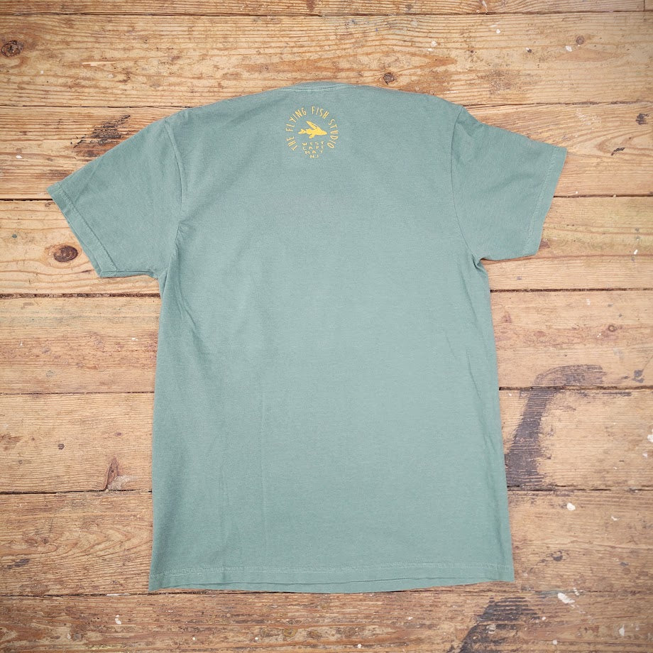 The back of a green T-shirt laid flat on a wooden surface, with the 'Flying Fish Studio' logo printed on the back of the neck in yellow ink.