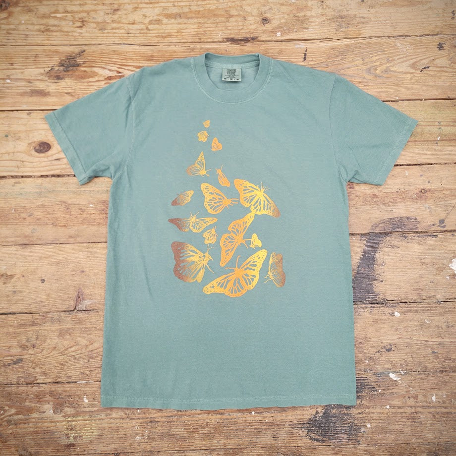 Green T-shirt with a graphic design of scattered butterflies on the front, printed with brown, yellow and orange ink. The shirt is laid flat on a wooden surface. 