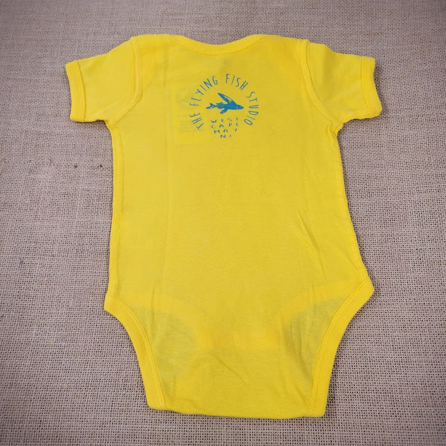 A bright yellow baby onesie featuring a cartoon flying fish. The text "THE FLYING FISH STUDIO, WEST CAPE MAY NJ" is printed below the design.