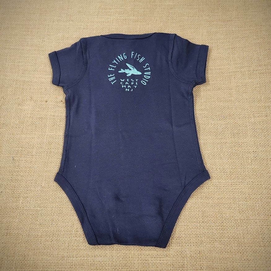 A navy baby onesie featuring a cartoon flying fish. The text "THE FLYING FISH STUDIO, WEST CAPE MAY NJ" is printed around & below the design.