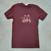 Maroon T-shirt  with a graphic design of two sloths riding a tandem bike laid flat on a blue textured surface.