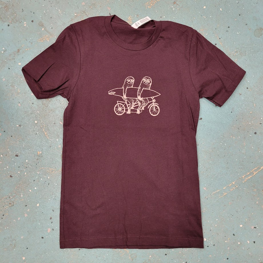 Maroon T-shirt  with a graphic design of two sloths riding a tandem bike laid flat on a blue textured surface.