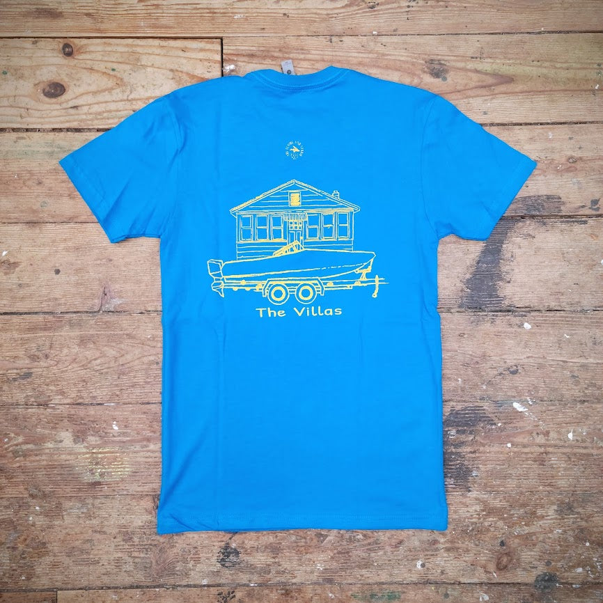 Blue T-shirt with a graphic design of a boat in front of a house and 'The Villas' printed on the back in yellow ink on a wooden surface.
