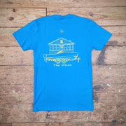 Blue T-shirt with a graphic design of a boat in front of a house and 'The Villas' printed on the back in yellow ink on a wooden surface.