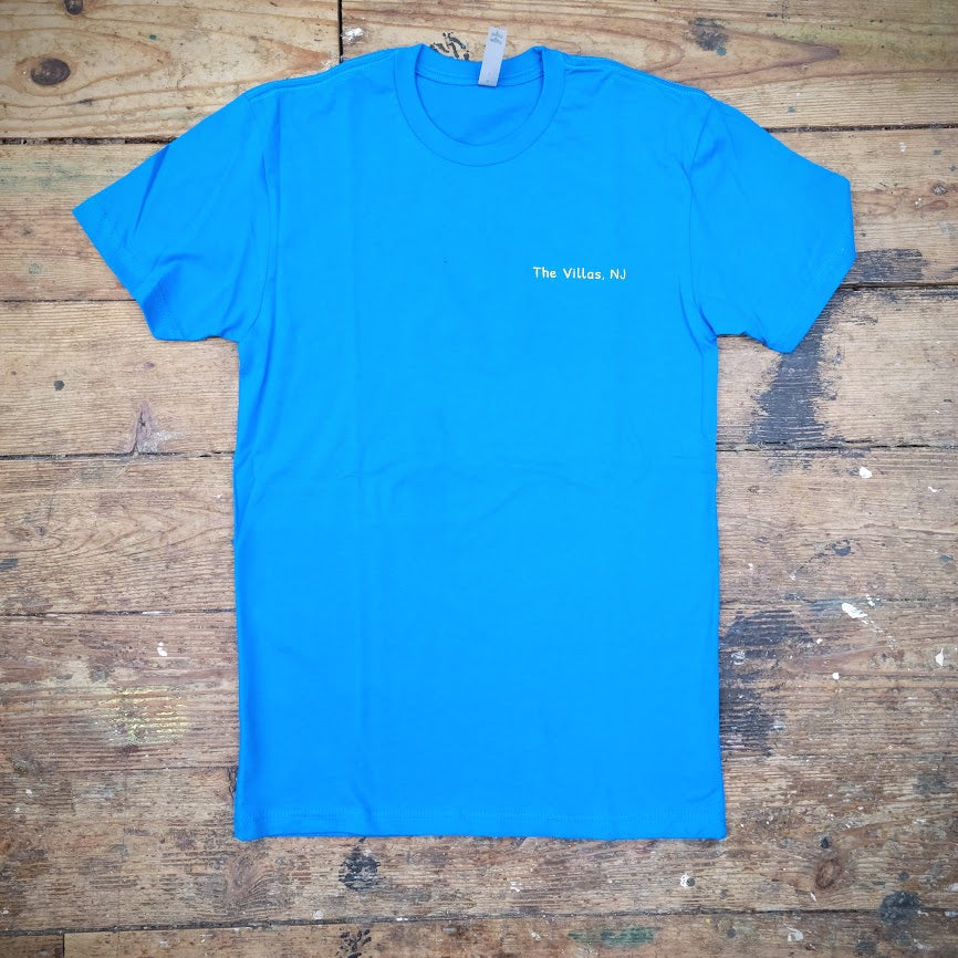 Blue T-shirt with 'The Villas, NJ' text printed on the left chest in yellow ink, laid flat on a wooden surface.