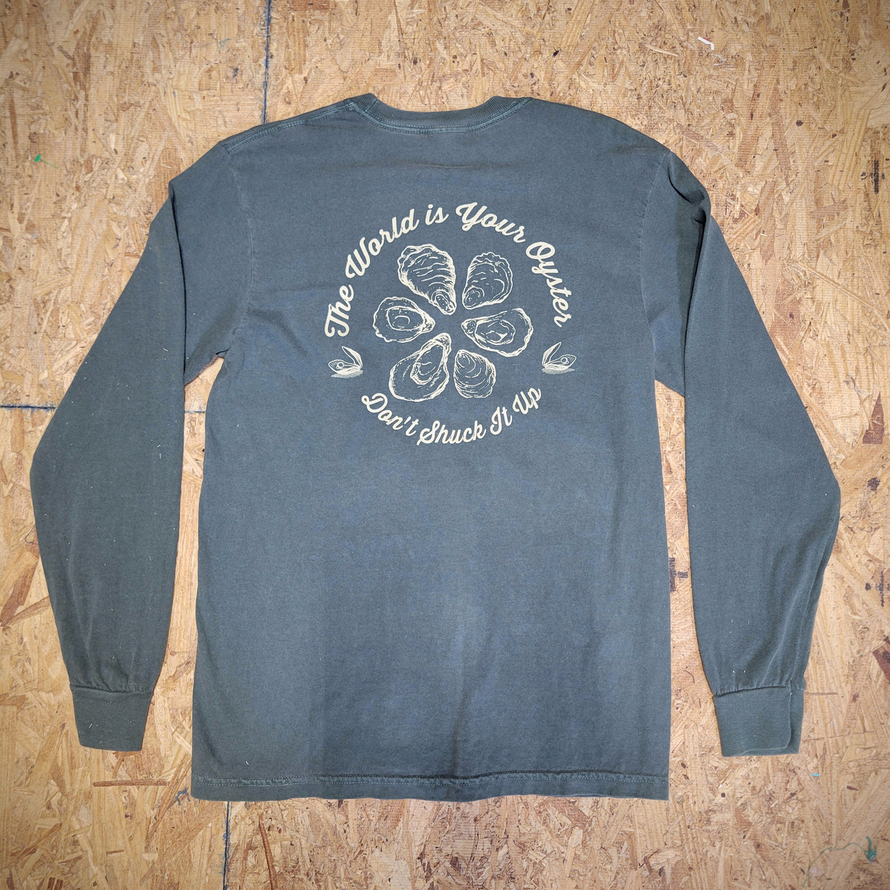 The back of a green long-sleeve T-shirt with a graphic design of oysters and the words, 'The World is Your Oyster. Don't shuck it up!' screen-printed in cream ink.