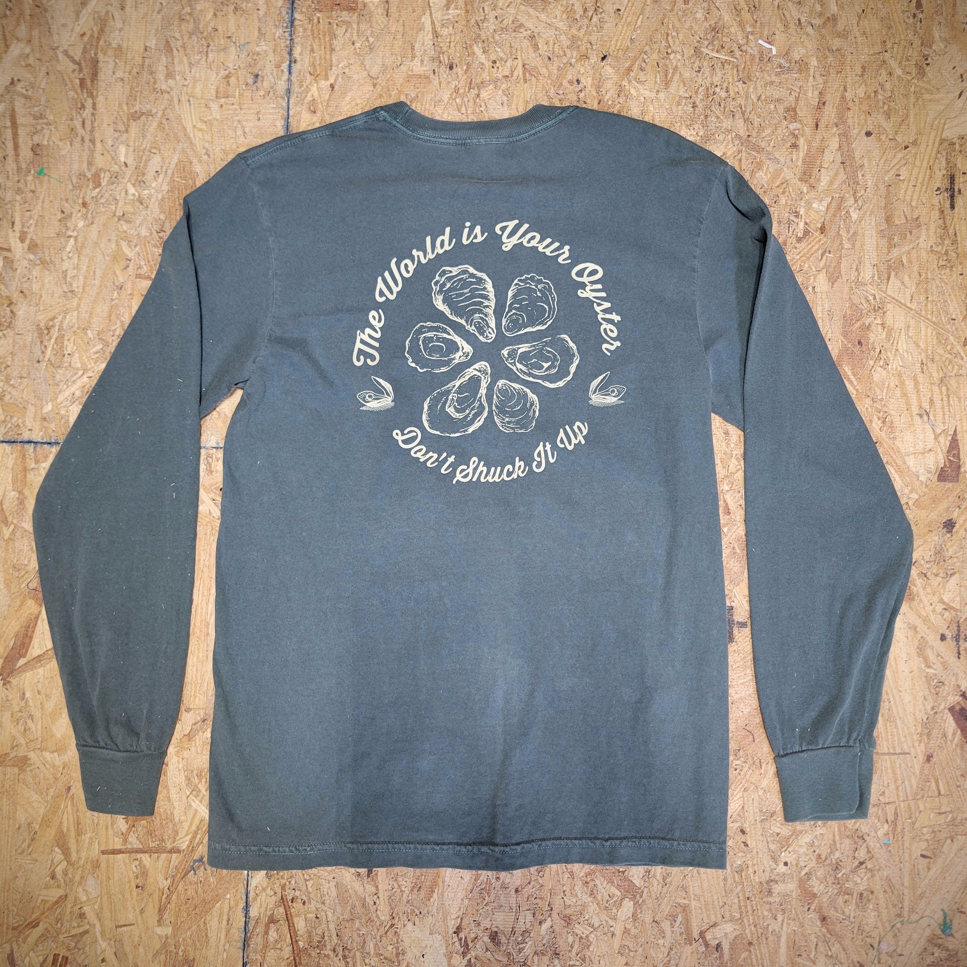 The back of a green long-sleeve T-shirt with a graphic design of oysters and the words, 'The World is Your Oyster. Don't shuck it up!' screen-printed in cream ink.
