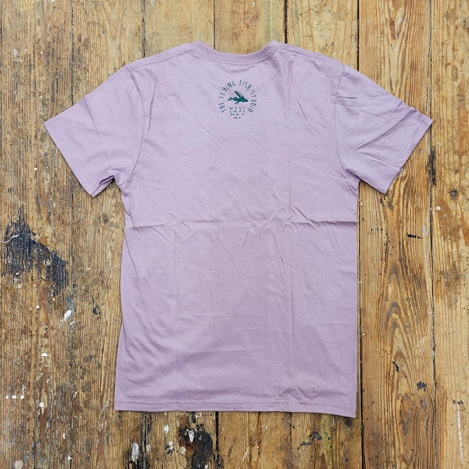 A light purple t-shirt is laid flat on a wooden surface, displaying its back. A green circular logo with "THE FLYING FISH STUDIO" encircles a stylized fish graphic in the upper back area of the shirt.