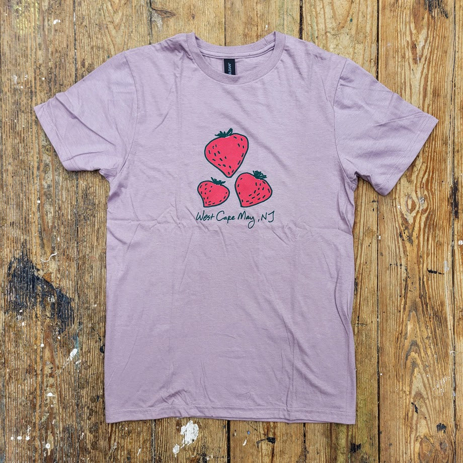 A light purple t-shirt lies flat on a wooden surface, featuring a graphic of three red strawberries of varying sizes. Below the strawberries, "West Cape May, NJ" is handwritten in a casual script.