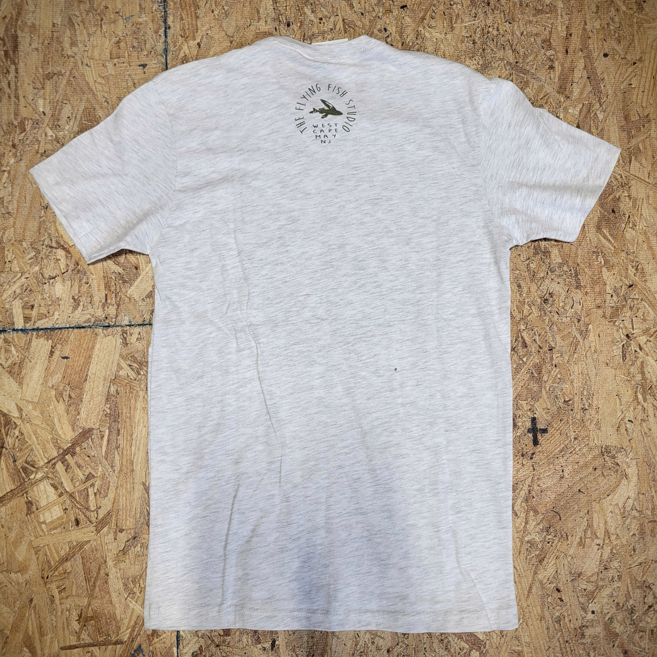Grey, heathered T-shirt laid flat on a wooden background with the 'Flying Fish Studio' logo on the back of the neck in green ink.