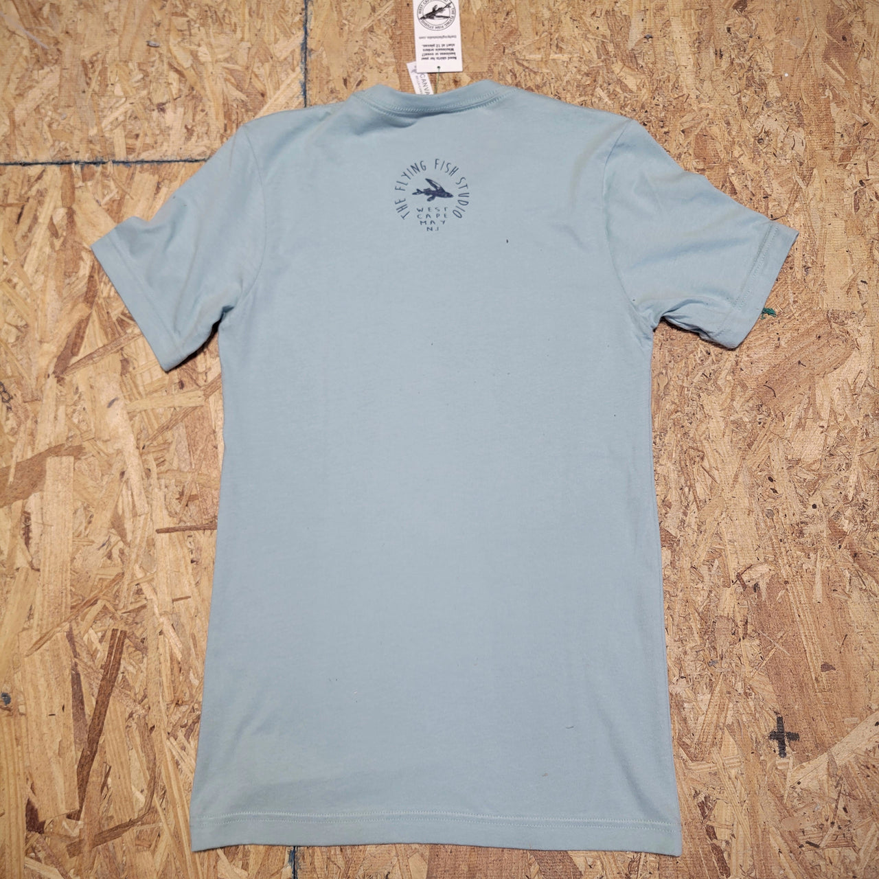 Back of a light blue, short sleeve T-shirt laid flat on a wooden surface. The 'Flying Fish Studio' logo is screen-printed on the back of the neck in dark blue ink. The words 'West Cape May, NJ' sit beneath it.