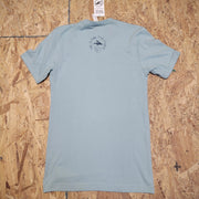 Back of a light blue, short sleeve T-shirt laid flat on a wooden surface. The 'Flying Fish Studio' logo is screen-printed on the back of the neck in dark blue ink. The words 'West Cape May, NJ' sit beneath it.