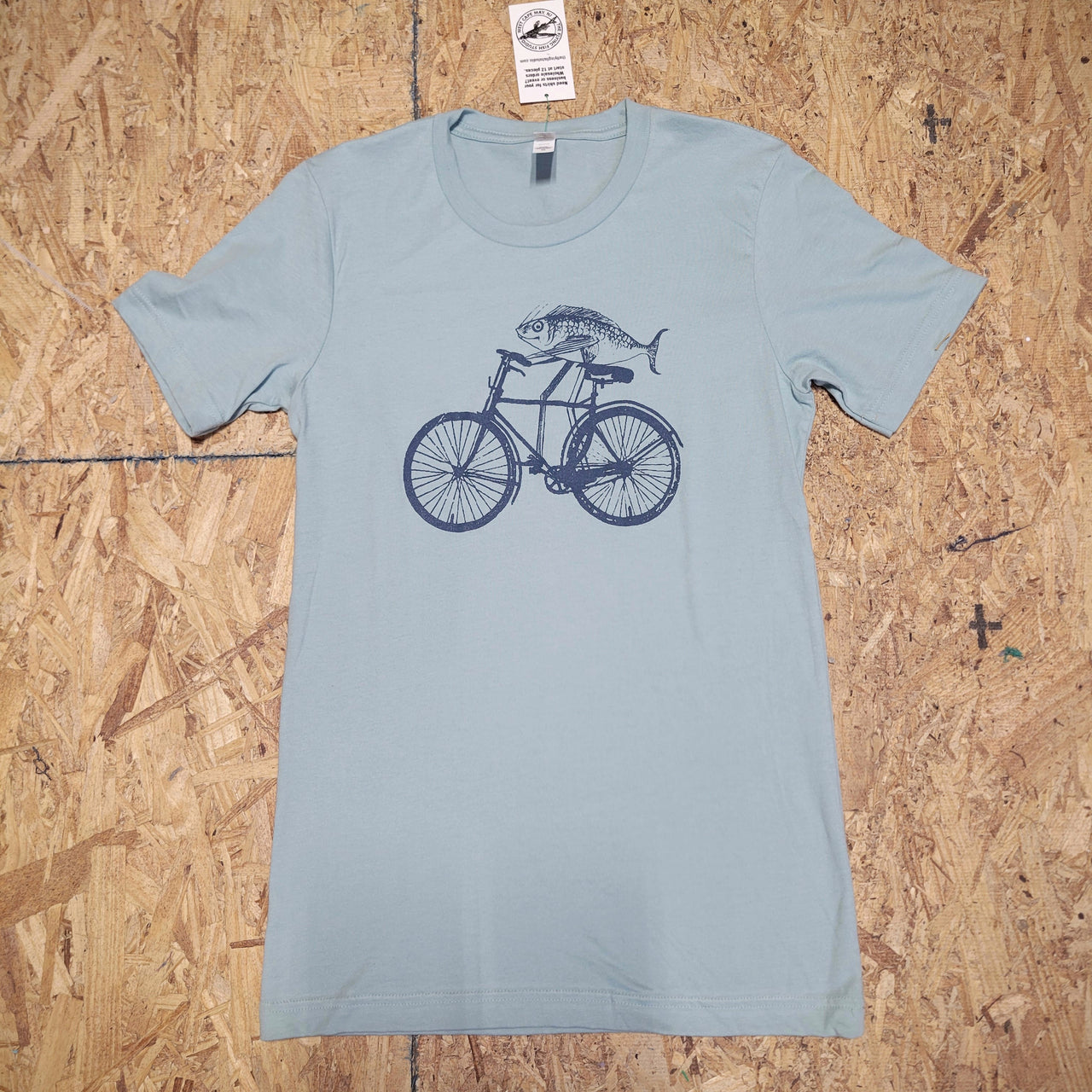 Front of a light blue, short sleeve T-shirt with a design of a fish riding a bike screen-printed on the front in dark blue ink. The shirt is laid flat on a wooden surface.