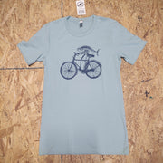 Front of a light blue, short sleeve T-shirt with a design of a fish riding a bike screen-printed on the front in dark blue ink. The shirt is laid flat on a wooden surface.
