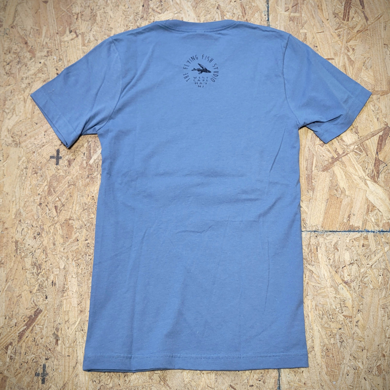The back of a blue T-shirt laid flat on a wooden surface with the 'Flying Fish Studio' logo screen-printed on the back of the neck in black ink.