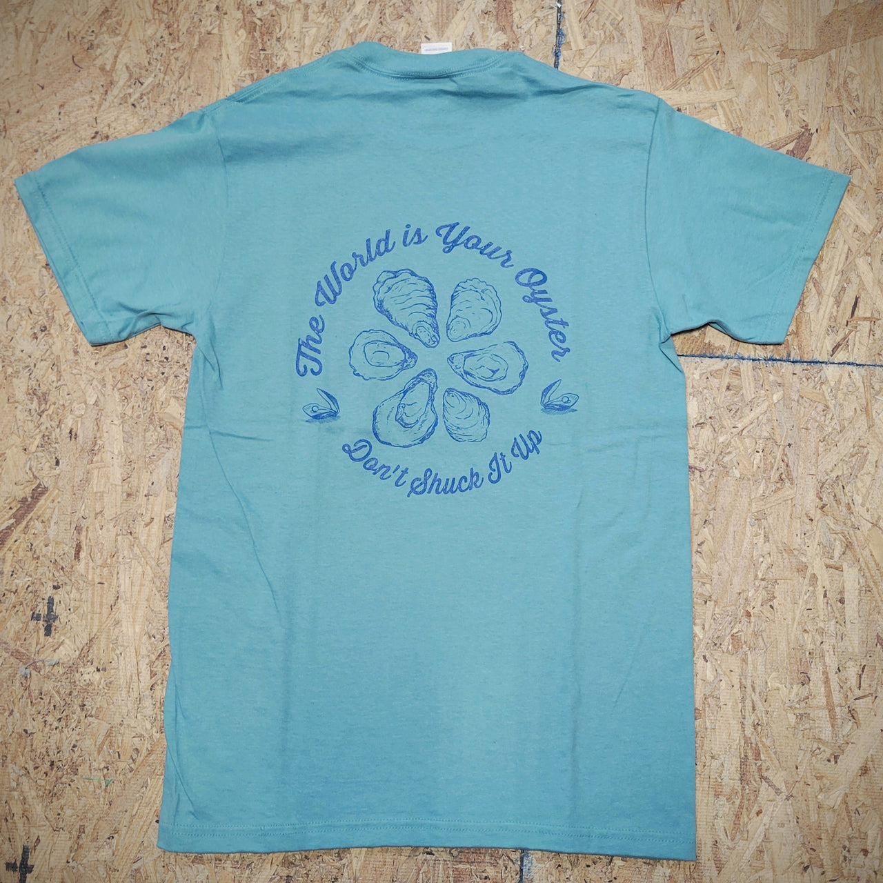 Greenish-blue T-shirt with a graphic design of oysters and text that says 'THE WORLD IS YOUR OYSTER! DON'T SHUCK IT UP!' on a wooden surface.