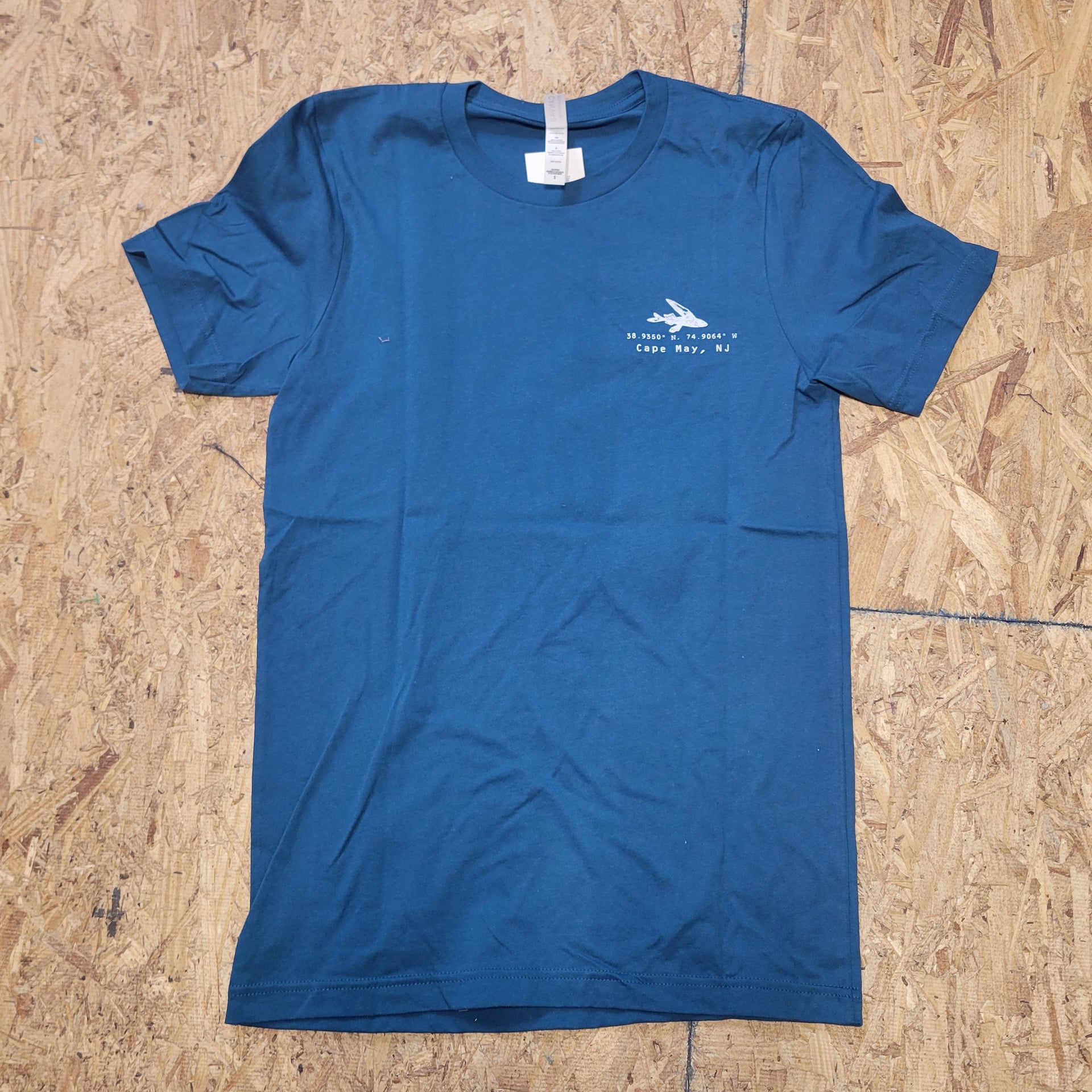 The front of a deep blue T-shirt laid flat on a wooden surface. The shirt has coordinates and a design of a fish on the left chest in white ink.
