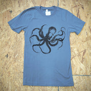 A blue T-shirt with a graphic design of an octopus screen-printed on the front in black ink. The shirt is laid flat on a wooden surface.