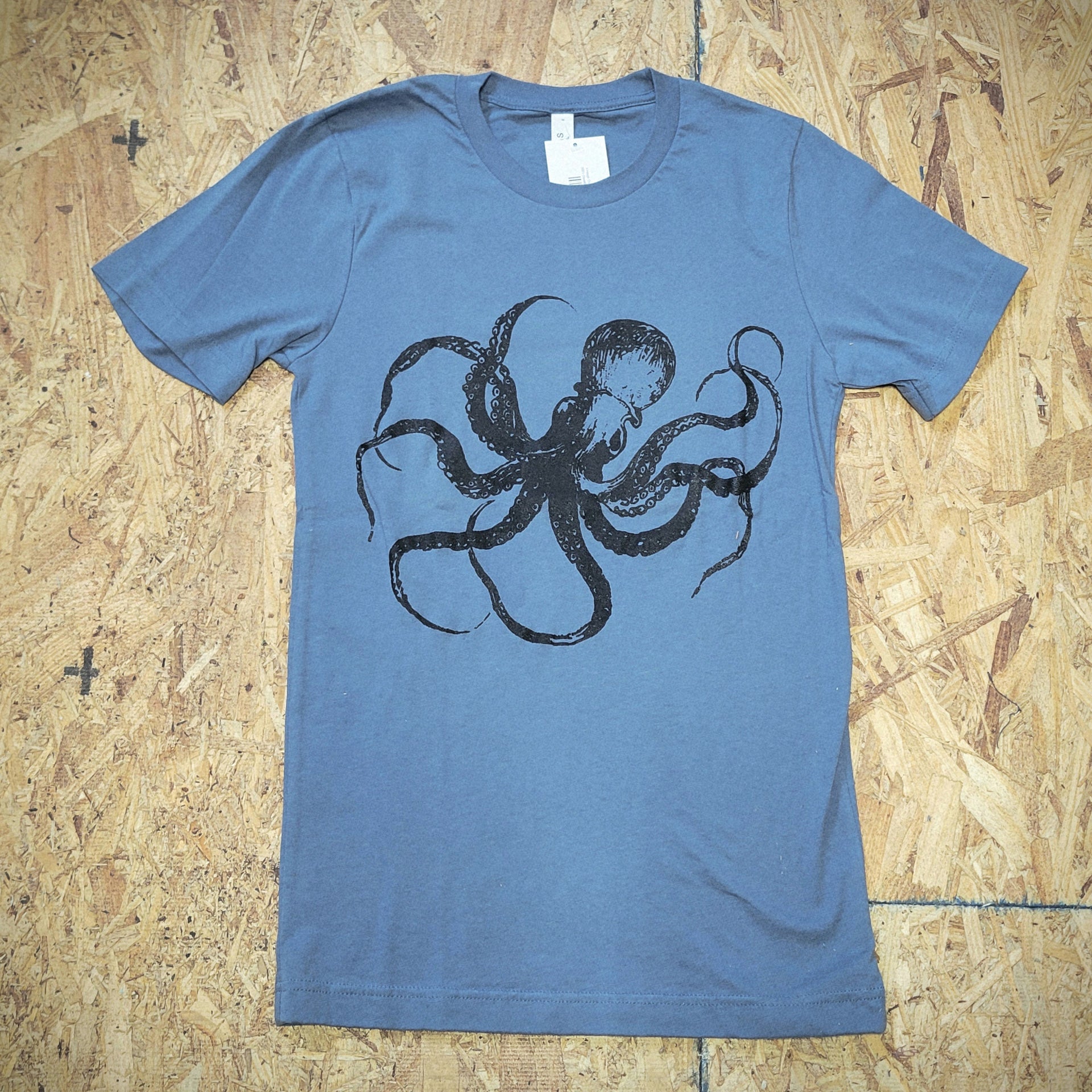 A blue T-shirt with a graphic design of an octopus screen-printed on the front in black ink. The shirt is laid flat on a wooden surface.