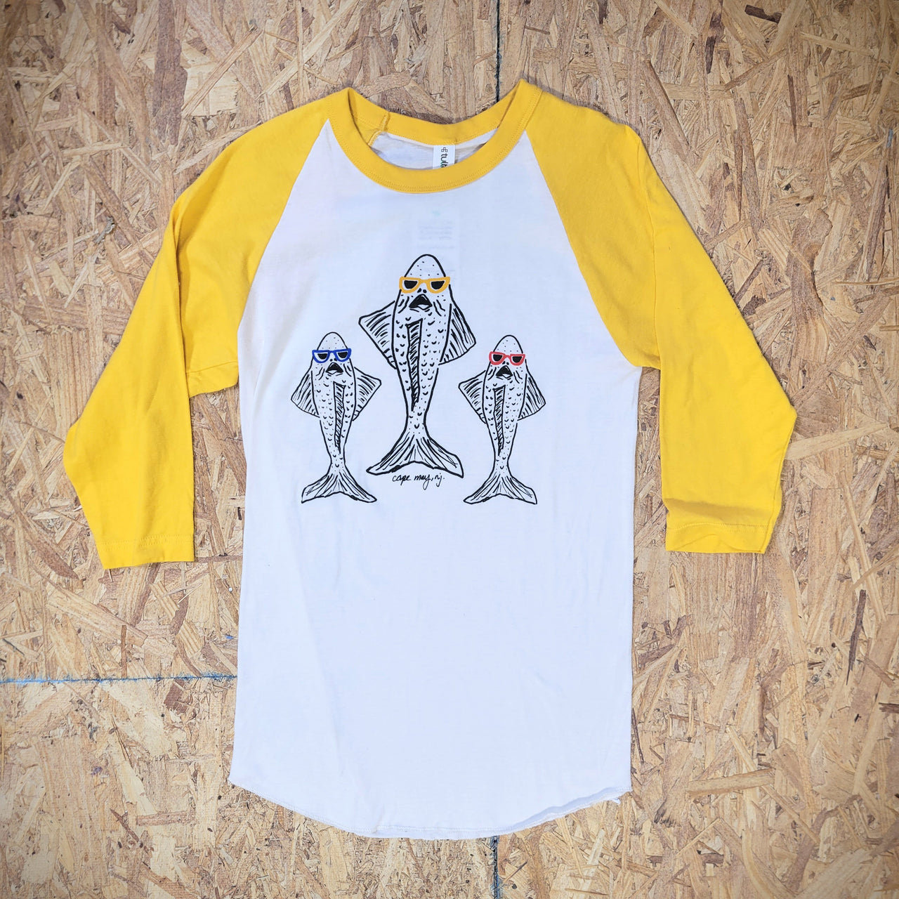 A yellow and white baseball T-shirt with a graphic design of three fish on  the front in black ink, wearing sunglasses in three different colors: blue, yellow, and red. The shirt is laid flat on a wooden surface and the words 'Cape May, NJ' are printed on the front underneath the fish.