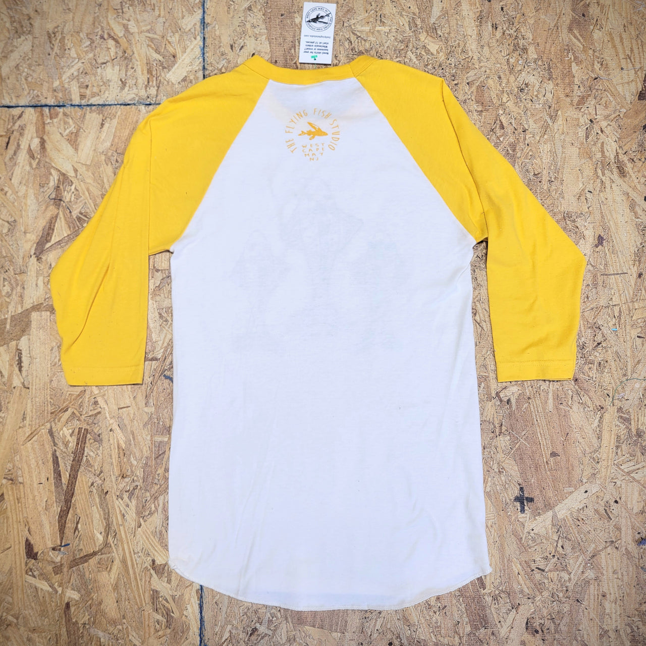 The back of a yellow and white baseball T-shirt laid flat on a wooden  surface. The 'Flying Fish Studio' logo is screen-printed on the back of the neck in yellow ink.