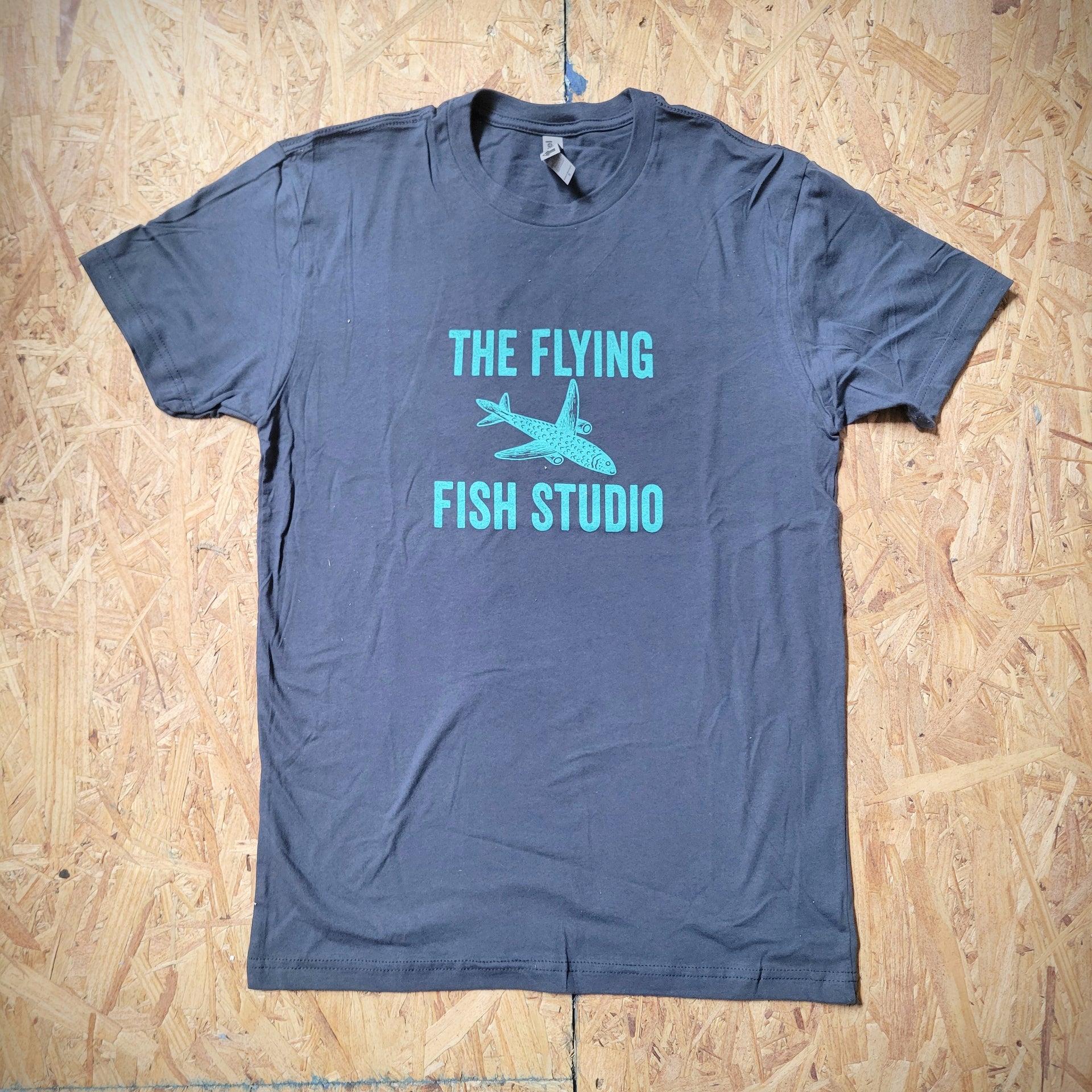 A blue T-shirt laid flat on a wooden background with a design of an airplane fish on the front and the words, "THE FLYING FISH STUDIO."