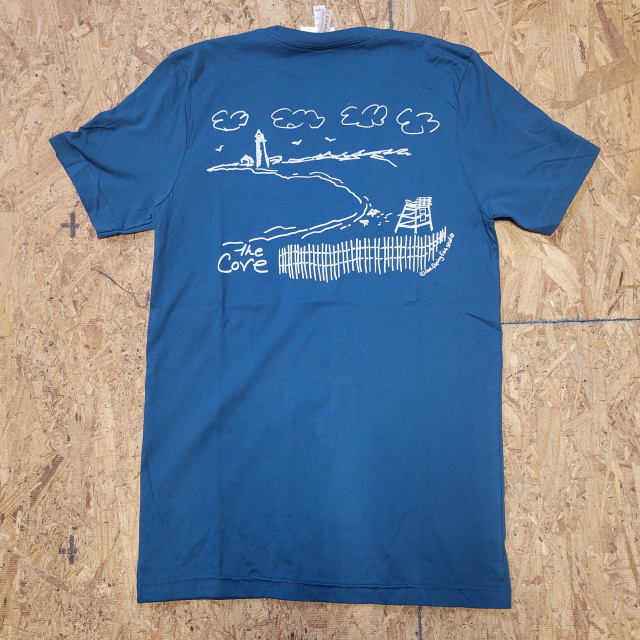 Image of a deep blue T-shirt with a white graphic design of a beach and text that says 'The Cove', laid flat on a wooden surface.