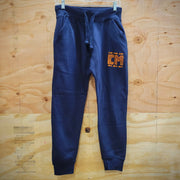 A pair of navy blue sweatpants hanging on a white hanger against a light wood panel background. The sweatpants feature a drawstring waist, side pockets, ribbed cuffs, and a stylized orange and yellow 'CM' graphic on the upper left thigh.