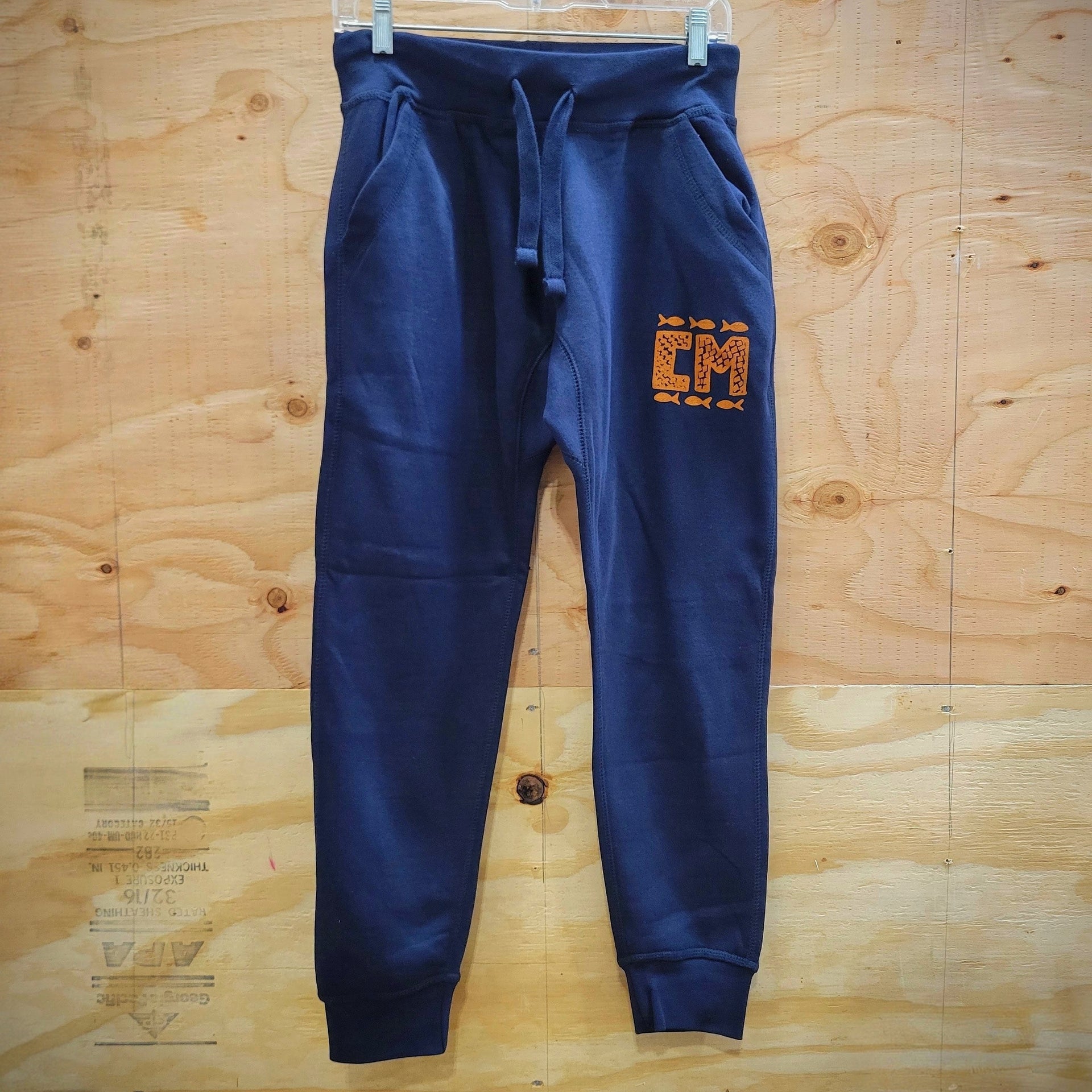 A pair of navy blue sweatpants hanging on a white hanger against a light wood panel background. The sweatpants feature a drawstring waist, side pockets, ribbed cuffs, and a stylized orange and yellow 'CM' graphic on the upper left thigh.