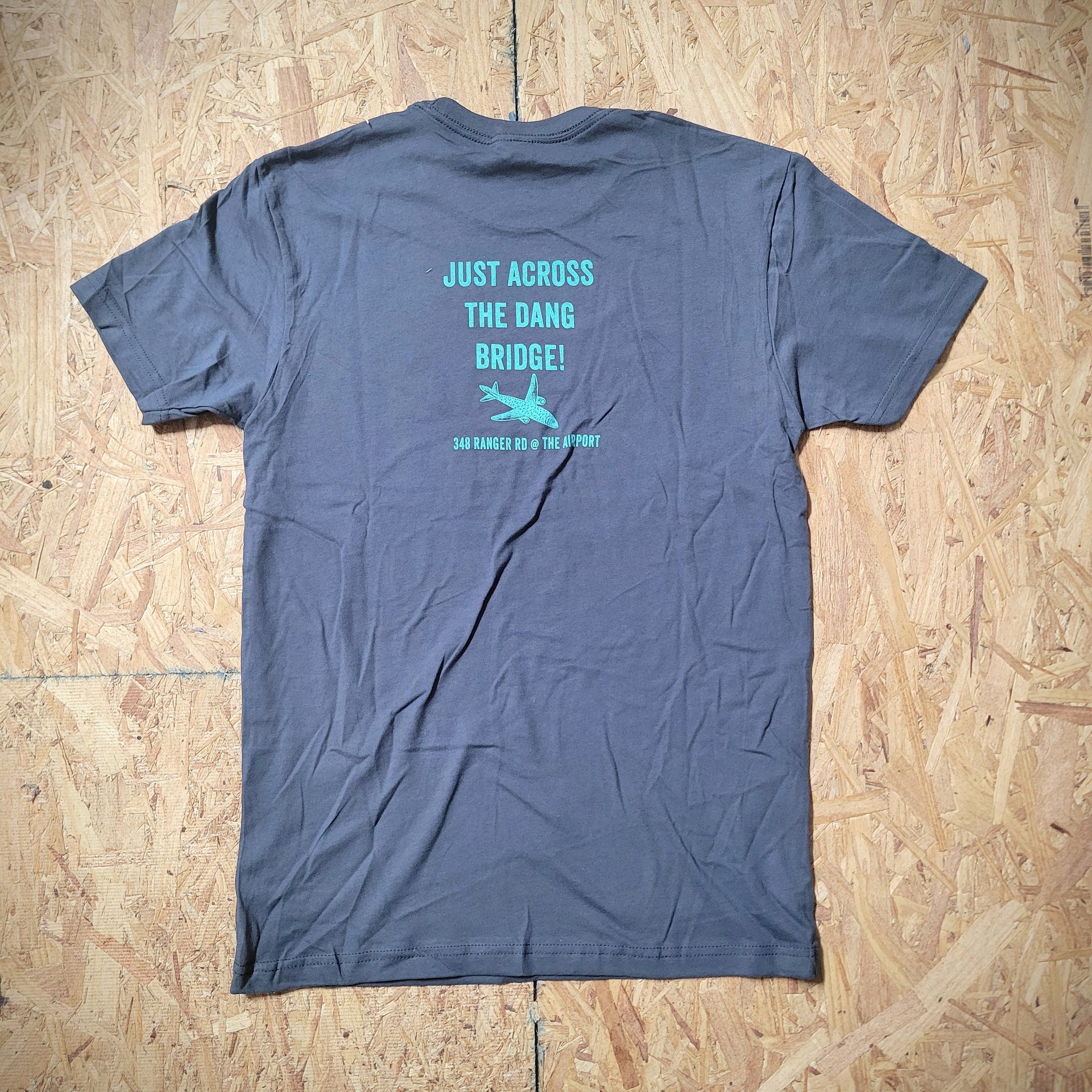 A blue T-shirt laid flat on a wooden surface with the words, "JUST ACROSS THE DANG BRIDGE."