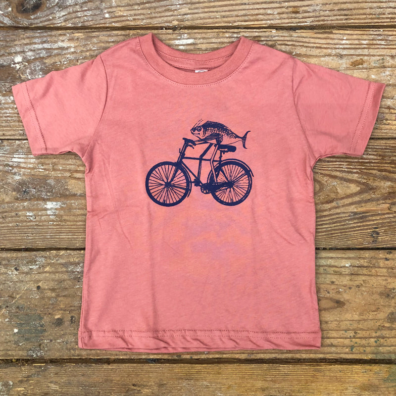 A mauve t-shirt featuring a navy-colored fish on a bike on the front chest.