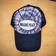 A black tie-dye trucker hat with an 'Imagine Peace' patch on it.
