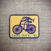 Rectangle, yellow, blue-rimmed patch that features, 'Cape May, NJ' and a fish riding a bike on the front.