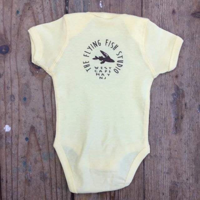Pastel yellow onesie featuring the 'Flying Fish' logo on the upper back in brown ink.